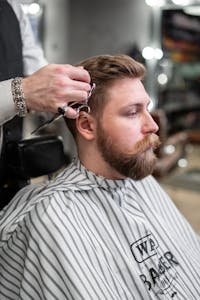 A barber trims a bearded man's hair in a classic barbershop setting.